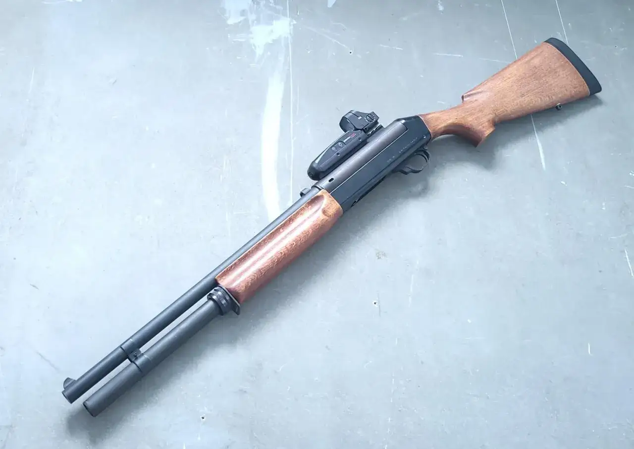 Fusil Benelli 121 avec viseur reflex, bois sombre et canon noir, pos&eacute; sur une surface grise.