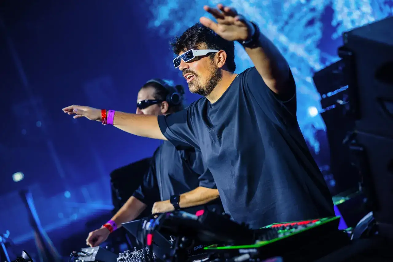 Dwóch DJ-ów, niczym agenci czasu, manipuluje dźwiękiem na scenie. Jeden w białych okularach, drugi w słuchawkach, obaj z uniesionymi rękami.