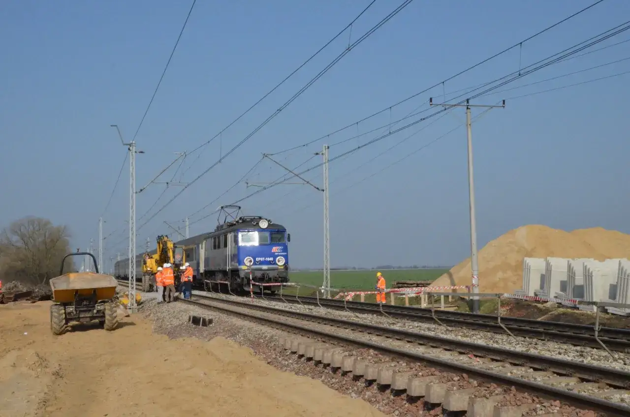 Prace torowe na trasie Oława-Wrocław. Lokomotywa EP07-1040 stoi na torach, obok koparka i wywrotka. Pracownicy w kamizelkach.