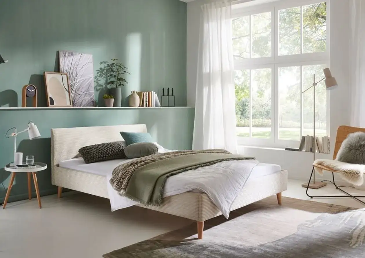Ein helles, skandinavisches schlafzimmer mit einem Bett, das mit einer grünen Decke und Kissen dekoriert ist. Ein großer Fenstersitz mit Blick auf einen Garten.
