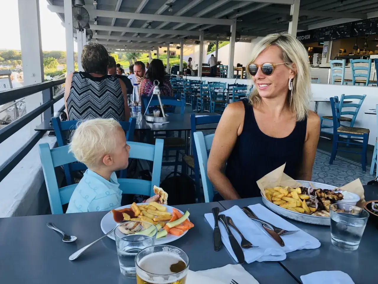Familj njuter av en m&aring;ltid p&aring; Costas Kitchen Kallithea. En kvinna ler, en pojke &auml;ter, och maten ser uts&ouml;kt ut.