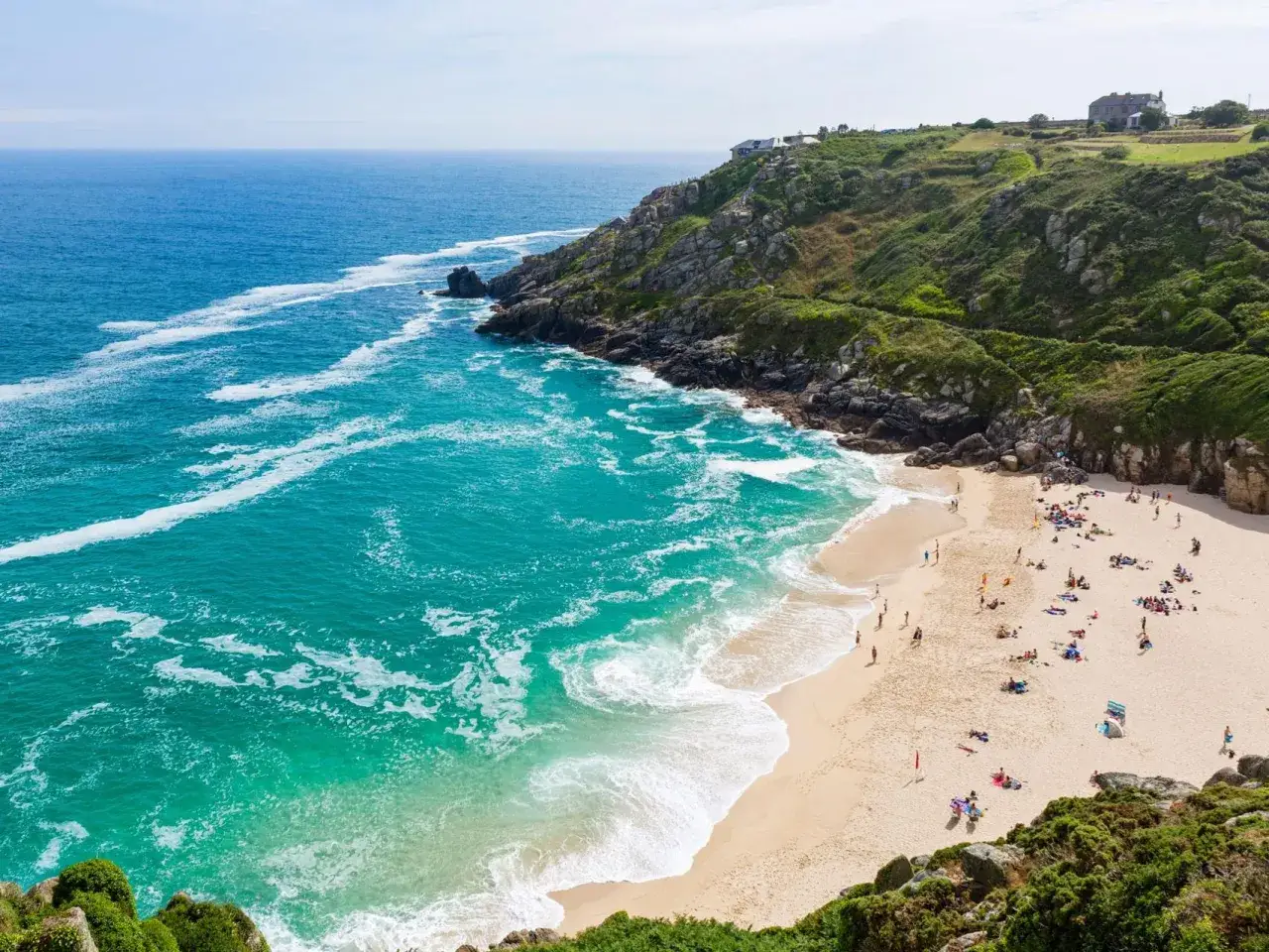 En solig dag vid St Ives. Turkosblått vatten sköljer in mot en gyllene sandstrand där människor njuter av solen. Grönskande klippor reser sig mot himlen.