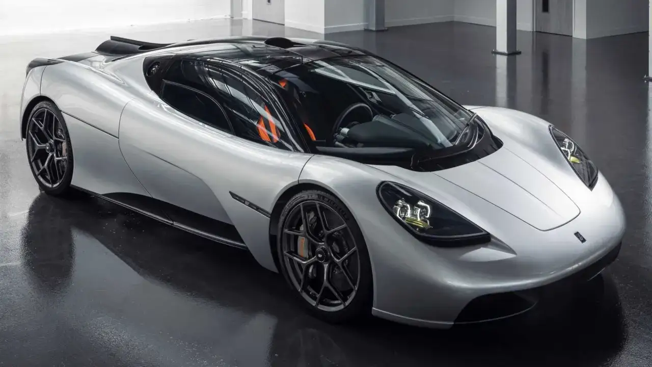 Der Gordon Murray T.50, ein silberner Supersportwagen mit orangefarbenen Akzenten im Innenraum, steht auf einem glänzenden Boden.