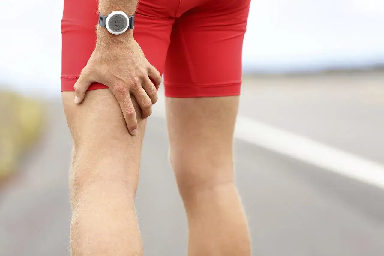 Un coureur porte un short rouge et une montre. Il se tient la cuisse, ressentant une douleur derrière la cuisse sans effort.