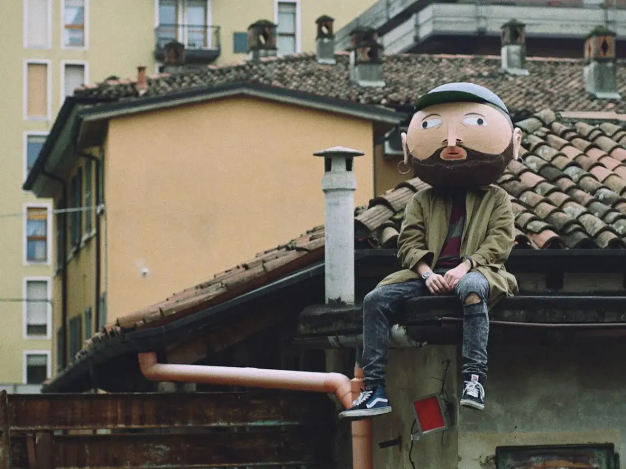 Un personaggio con una maschera gigante, simile a un cratere, siede sui tetti. I testi di Frah Quintale potrebbero raccontare storie di solitudine urbana.