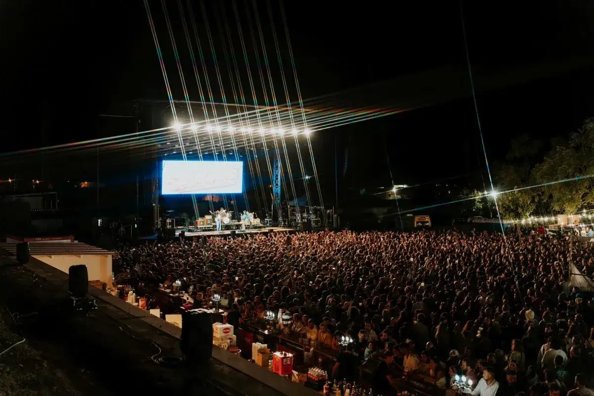 Multitud vibrante en un **amante festival** de música, con una banda tocando en el escenario iluminado y una gran pantalla de fondo.