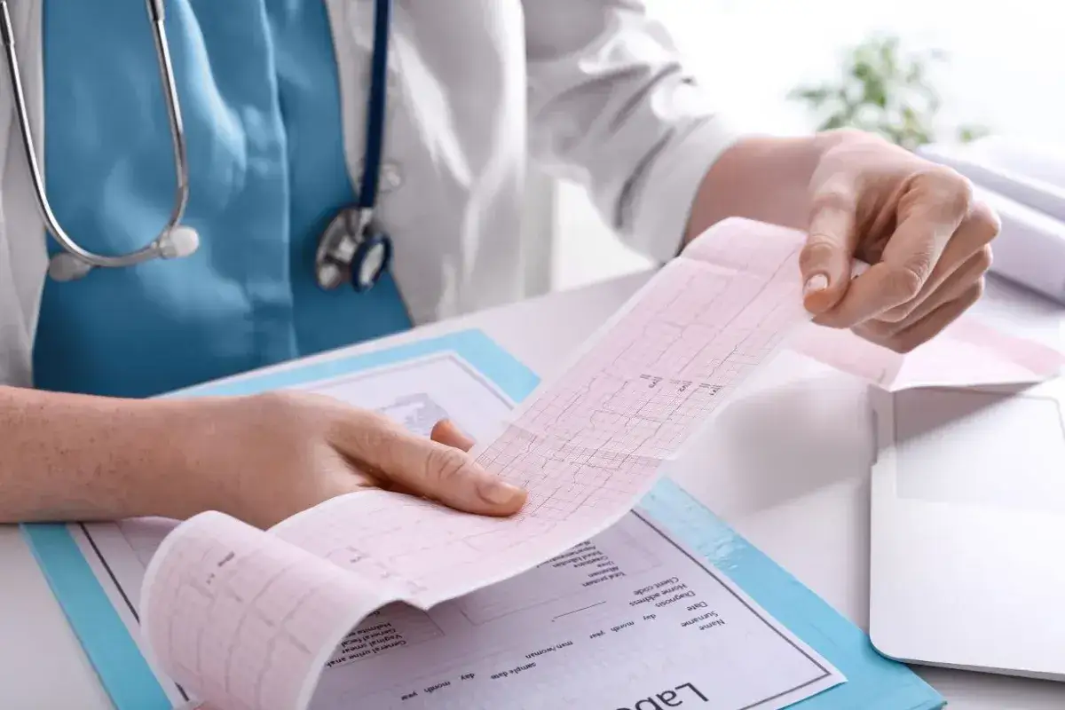 Pielęgniarstwo kardiologiczne: analizowanie EKG. Dłoń trzyma wydruk z zapisem pracy serca, obok dokumentacja medyczna.