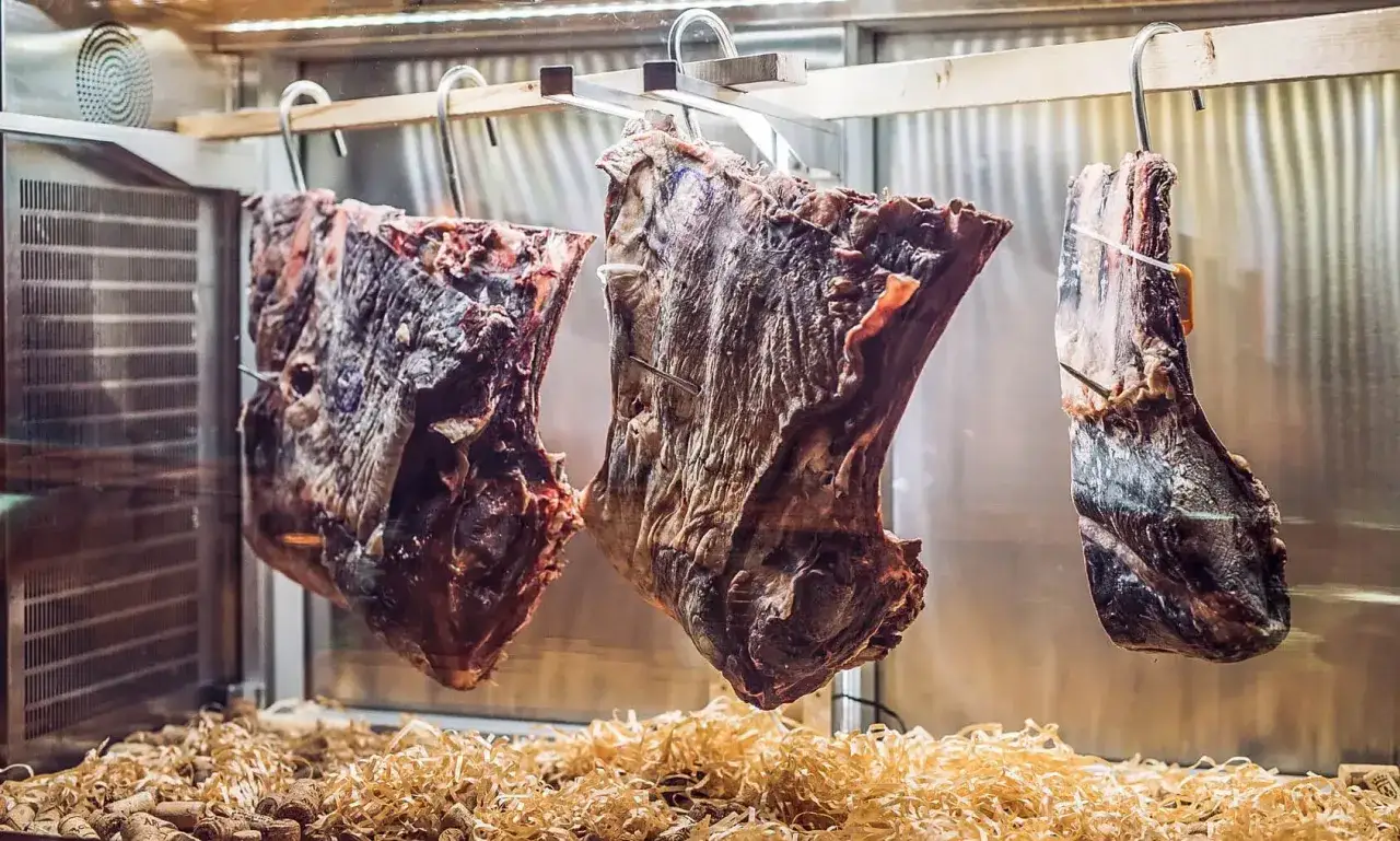 Drei große Stücke **rindfleisch abgehangen** reifen in einer Kühlkammer. Sie hängen an Haken über einer Unterlage aus Holzspänen und Korken.
