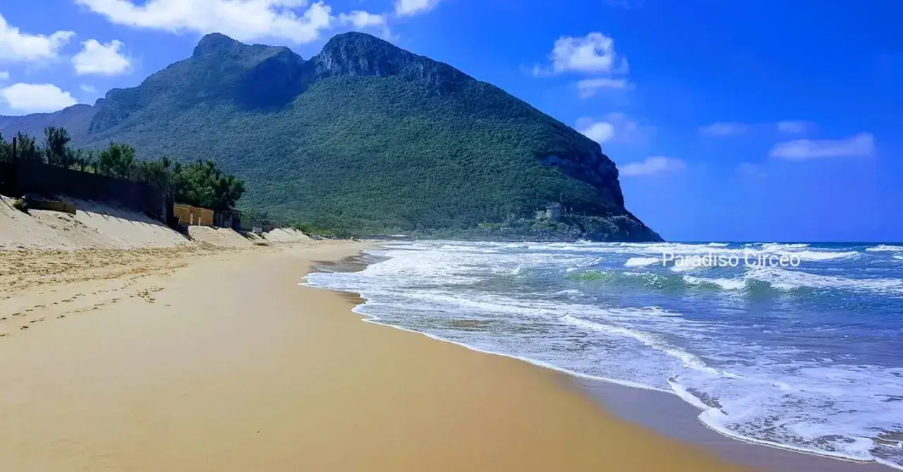 Spiaggia dorata e mare mosso sul litorale laziale, con il Monte Circeo verdeggiante sullo sfondo.