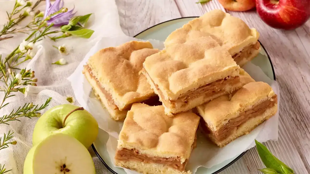 Saftiger gedeckter Apfelkuchen, in Quadrate geschnitten, liegt auf einem Teller. Frische Äpfel und zarte Blüten schmücken die Szene.