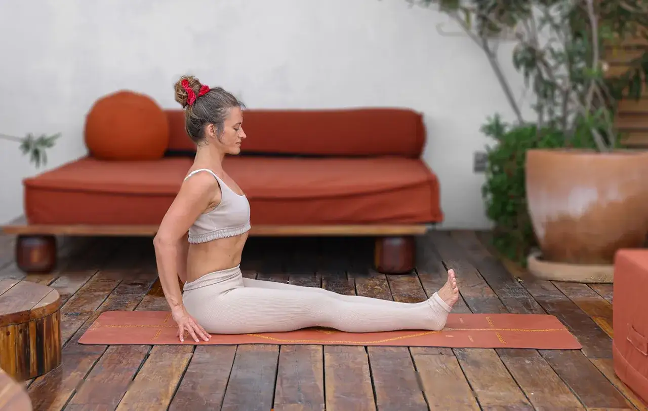 Mujer en postura sentada yoga, con piernas extendidas y manos apoyadas en el suelo detr&aacute;s de ella.