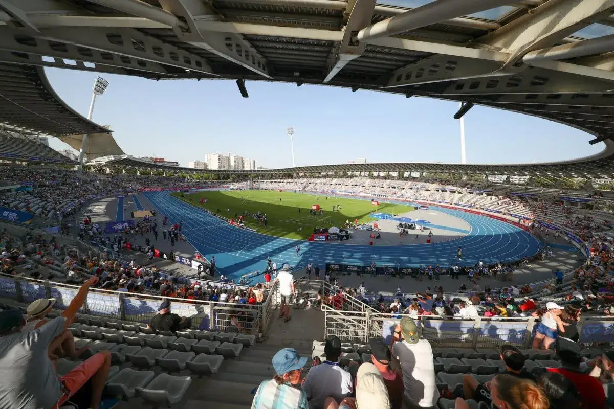 Vue panoramique du stade Charléty, avec une piste d'athlétisme bleue, un terrain de football vert et des gradins remplis de spectateurs sous un ciel bleu.