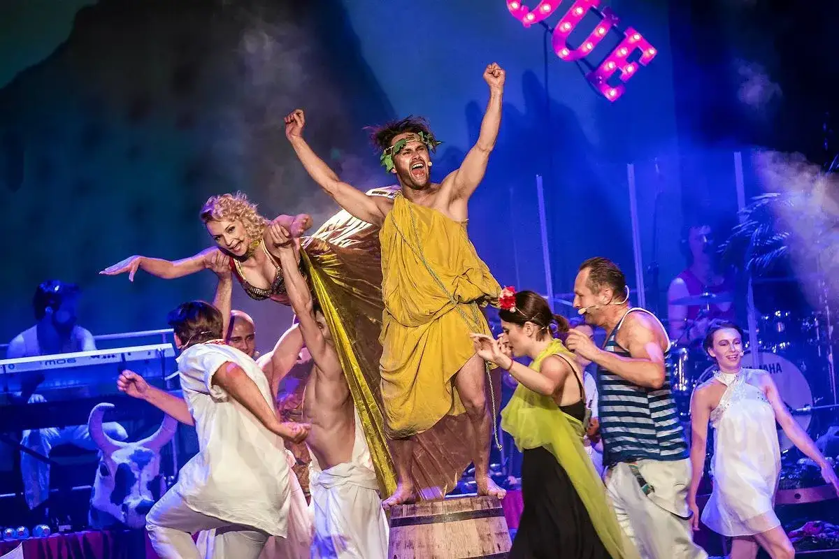Scena teatralna z tańczącymi ludźmi w strojach antycznych. W centrum mężczyzna w żółtej todze, z wieńcem laurowym na głowie, wznosi ręce. Obok niego kobieta w złotym stroju. W tle muzycy i neonowy napis 