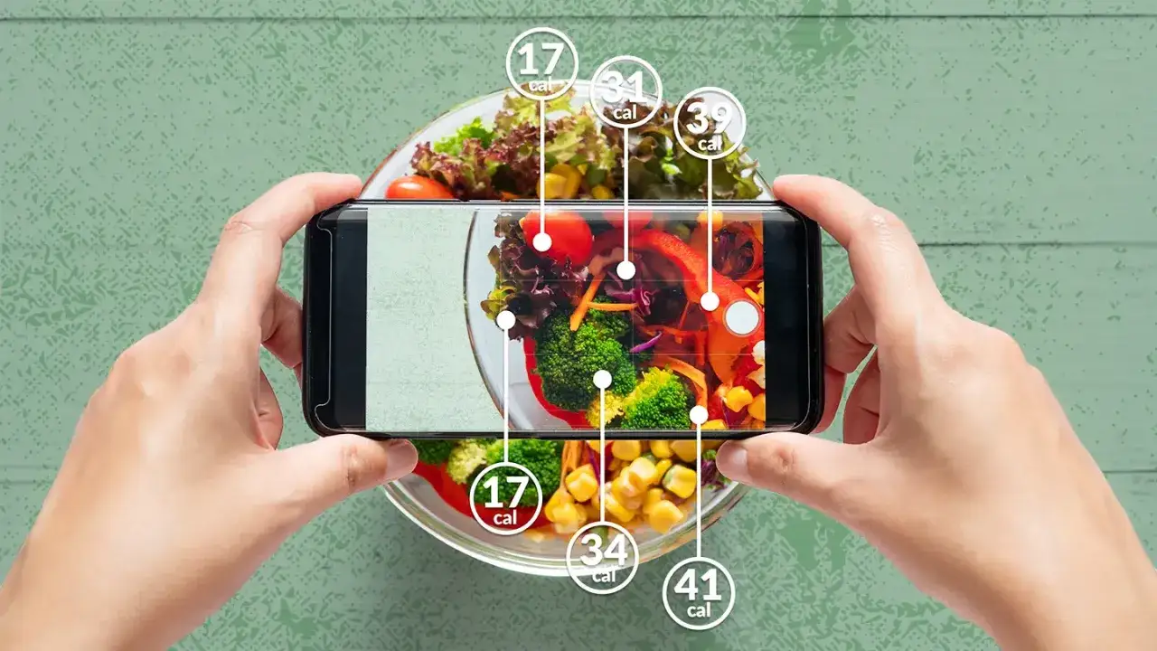 Ręce trzymają smartfon, który pokazuje aplikację do liczenia kalorii. Obrazek ilustruje, jak obliczyć deficyt kaloryczny, analizując składniki sałatki.