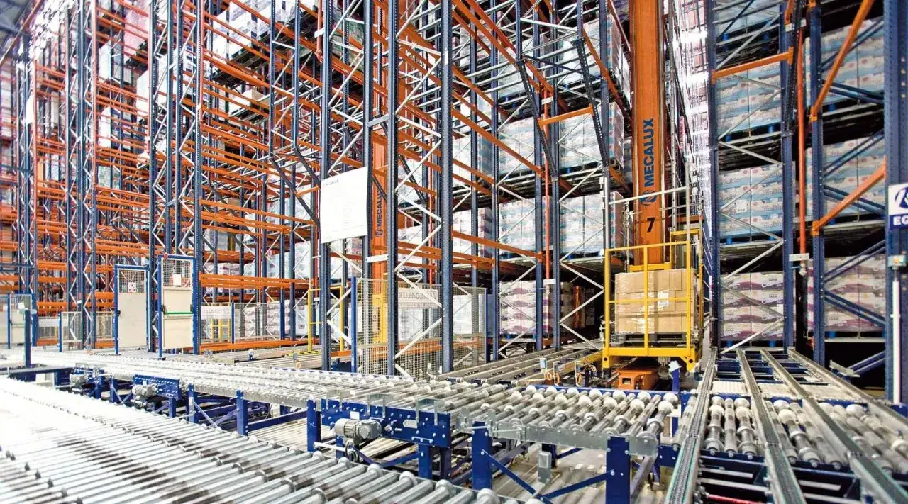 Automatyczny **stacker crane** porusza się po torach w magazynie wysokiego składowania, przenosząc palety z towarem między regałami.