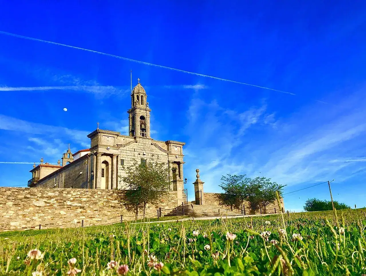Iglesia de piedra con campanario alto bajo un cielo azul vibrante. El corpiño de la iglesia se alza sobre un muro de piedra, con flores silvestres en primer plano.