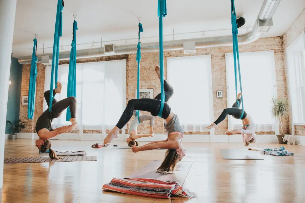 Kvinnor utf&ouml;r flying yoga i bl&aring;a tyger. De h&auml;nger upp och ner i olika positioner, en del med h&auml;nderna sammanfl&auml;tade.
