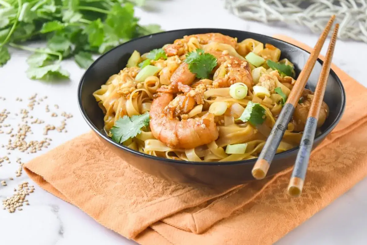 Un tazón de fideos de arroz con gambas, adornado con cilantro y cebolleta, listo para ser disfrutado con palillos.