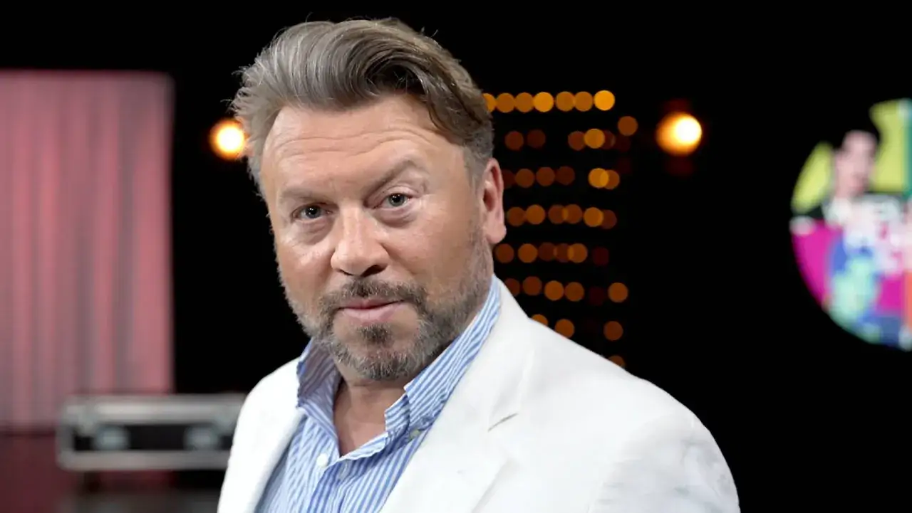 Michał Milowicz w białej marynarce, znany z filmów, seriali i programów, pozuje na tle świateł.