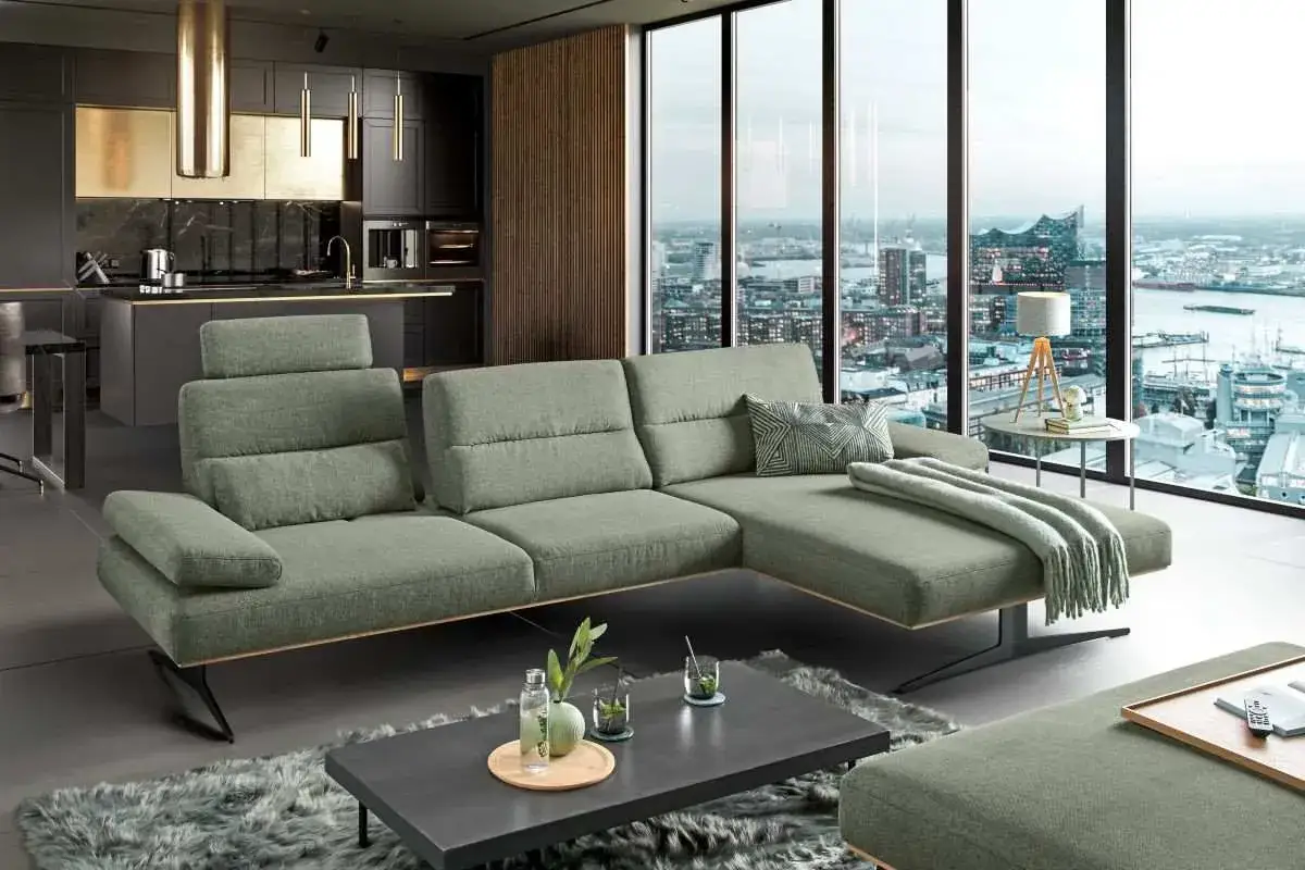 Ein modernes Wohnzimmer mit einem gro&szlig;z&uuml;gigen Ecksofa in sanfter Sofa Farbe, das sich vor einem Panoramafenster mit Stadtblick erstreckt.