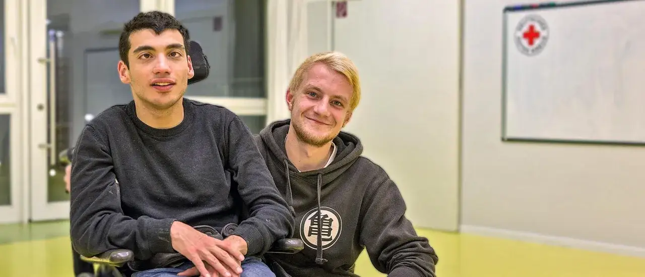 Zwei junge M&auml;nner, einer im Rollstuhl, l&auml;cheln in die Kamera. Sie sind ein Beispiel daf&uuml;r, was soziale Dienstleistungen bewirken k&ouml;nnen: Gemeinschaft und Unterst&uuml;tzung.