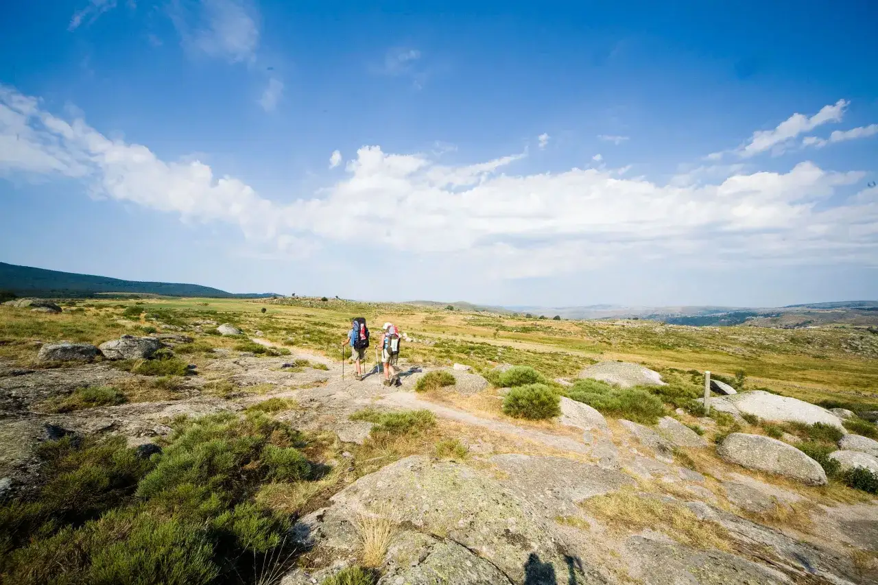 Deux randonneurs avancent sur un sentier rocailleux sous un ciel bleu. Le paysage est vaste, avec des collines verdoyantes et des rochers éparpillés. Le GR 70 offre une belle perspective.