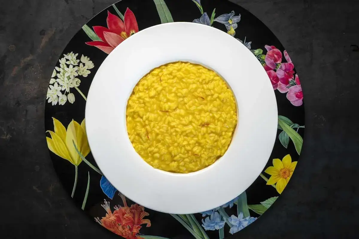 Un invitante risotto allo zafferano, dal colore dorato intenso, servito in una ciotola bianca su un piatto decorato con fiori colorati.