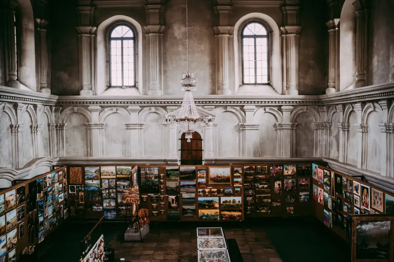 Wnętrze dawnej synagogi zamienionej na galerię sztuki. Obrazy wiszą na ścianach, a pośrodku wisi kryształowy żyrandol.