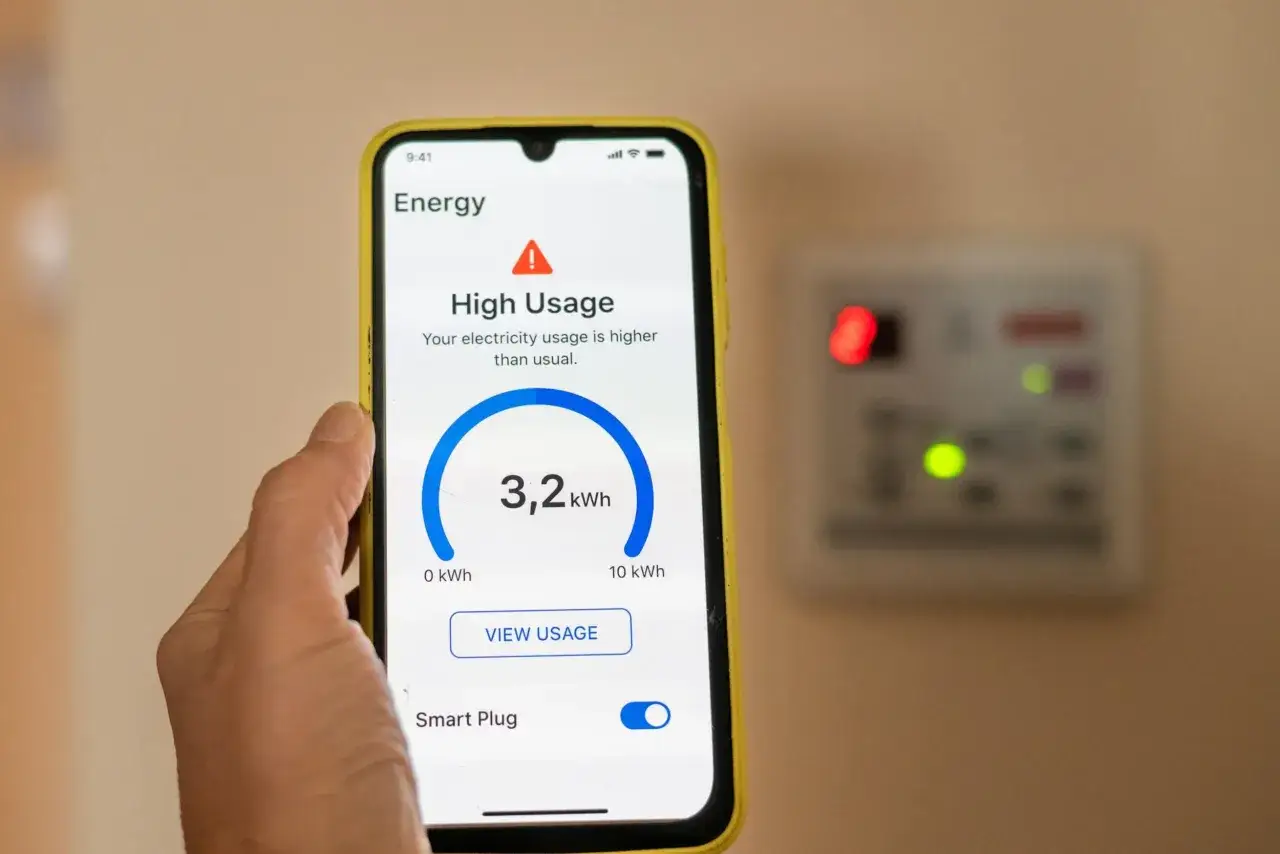 Teléfono muestra alto consumo eléctrico, 3,2 kWh. Detrás, un enchufe inteligente con luces indicadoras, parte de los sensores eléctricos.