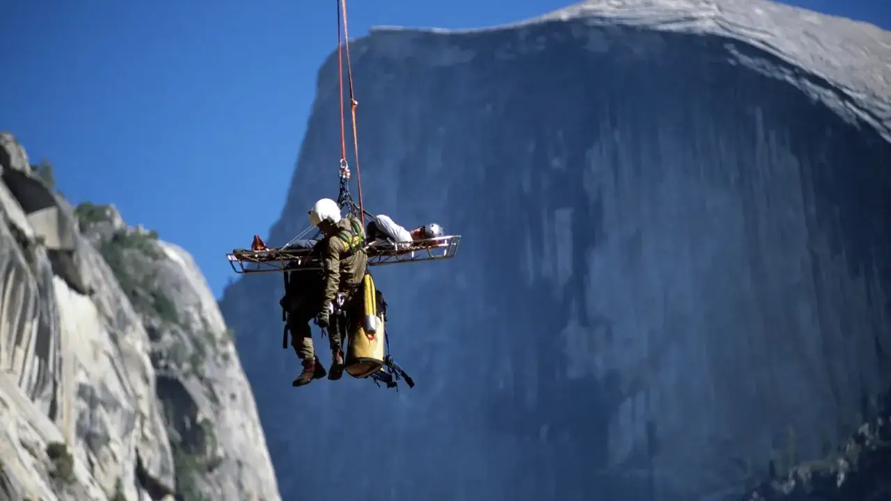 Rescuer in a helicopter hoist transports an injured climber, suspended in a stretcher, away from a sheer cliff face. This is what it means to be cliffed out: stuck on a cliff with no easy way down.