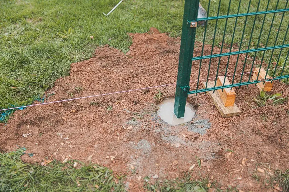 Ein grüner Zaunpfosten wird gerade mit Beton im Erdreich befestigt. Der Beton ist noch feucht und bildet einen Kreis um den Pfosten.