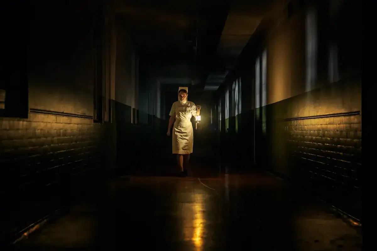 Mroczny korytarz szpitalny, pielęgniarka z latarnią w ręku. Atmosfera budzi niepokój, zapowiada thriller.