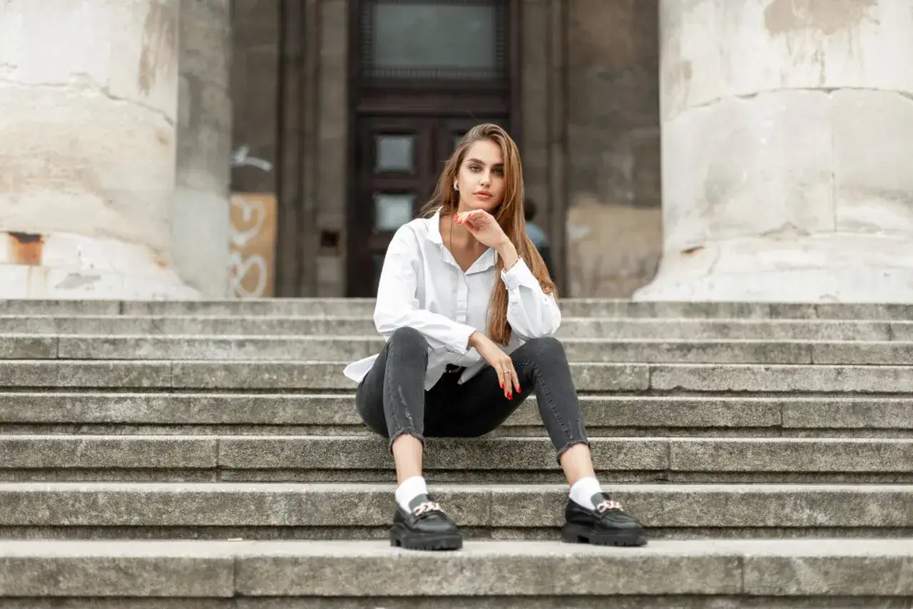 Eleganckie mokasyny to idealne buty do szarych spodni. Kobieta w białej koszuli i czarnych jeansach siedzi na schodach.