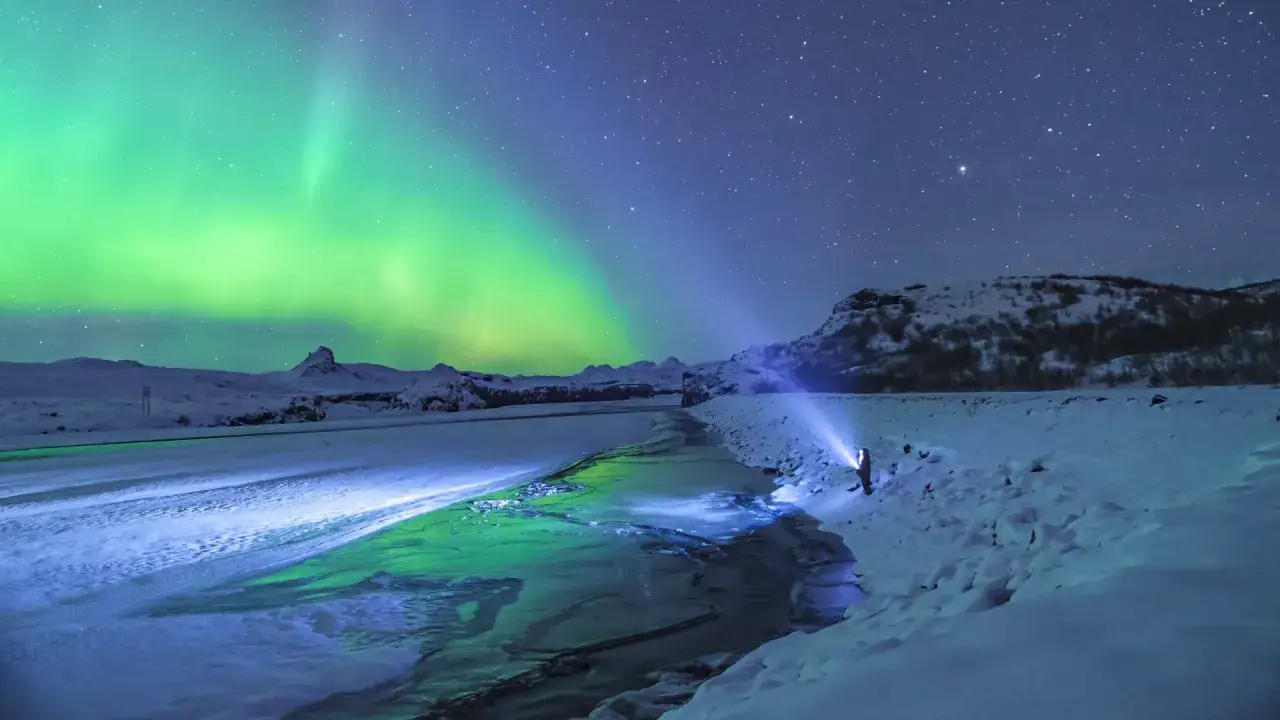 Lodowa rzeka i zorza polarna na nocnym niebie. Islandia w styczniu, człowiek z latarką oświetla brzeg.