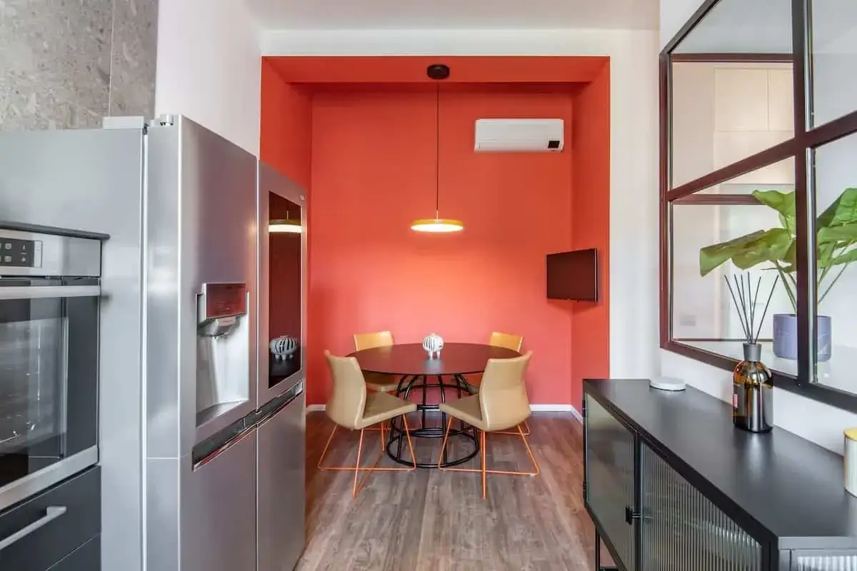Accogliente cucina in mansarda bassa con pareti corallo, tavolo rotondo e sedie color cuoio. Frigorifero in acciaio inox e un tocco di verde da una pianta.