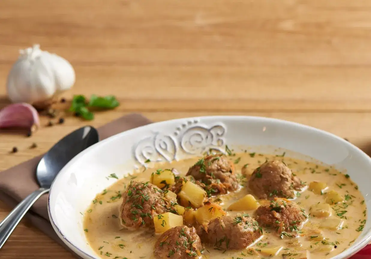 Zupa klopsowa babci z ziemniakami i koperkiem, podana w białej misce. Obok czosnek i przyprawy.