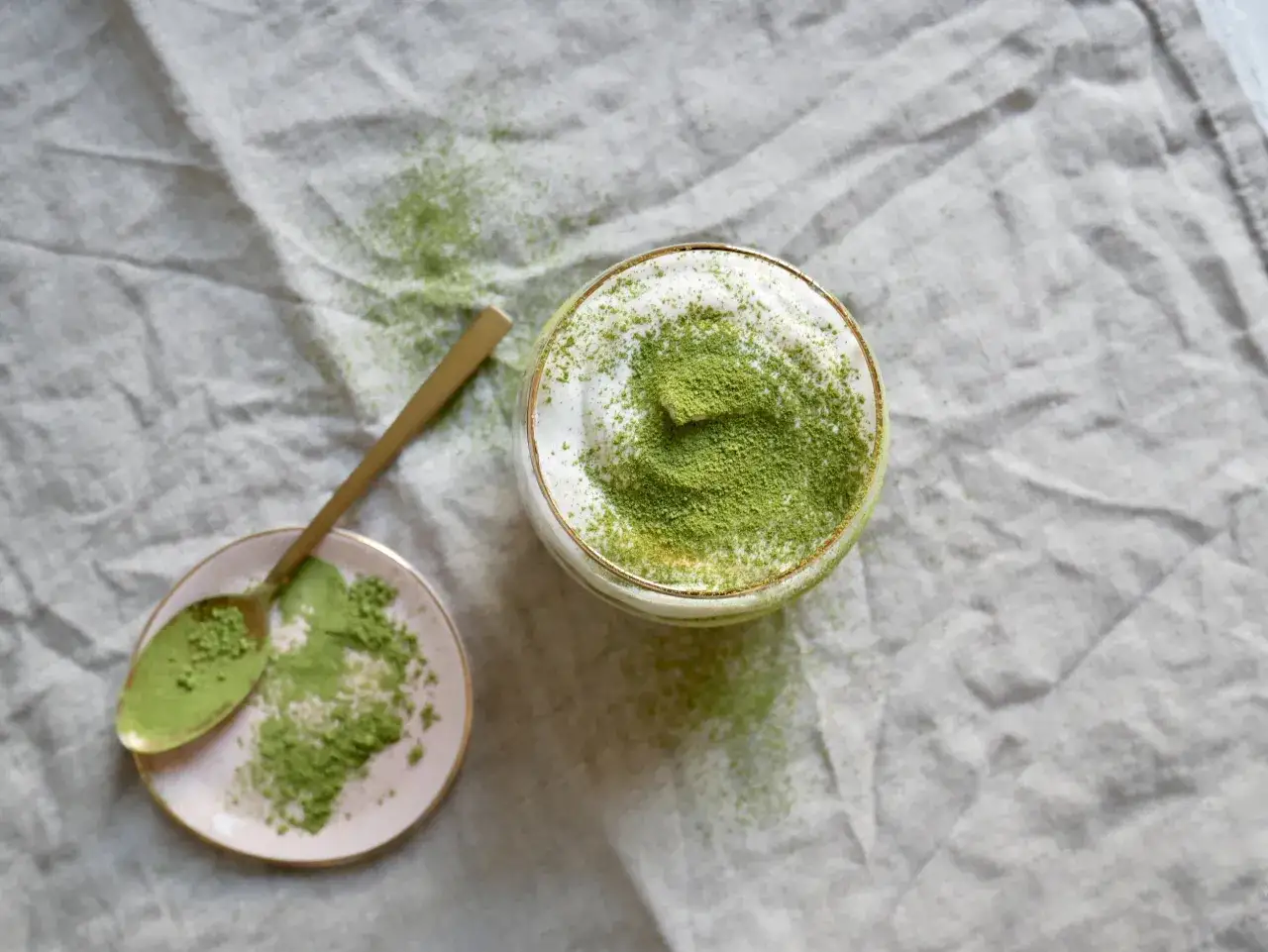 Ein Glas mit cremigem Matcha Tiramisu, bestäubt mit feinem Matcha-Pulver. Daneben ein Löffel auf einer kleinen rosa Schale, ebenfalls mit Matcha bestreut.