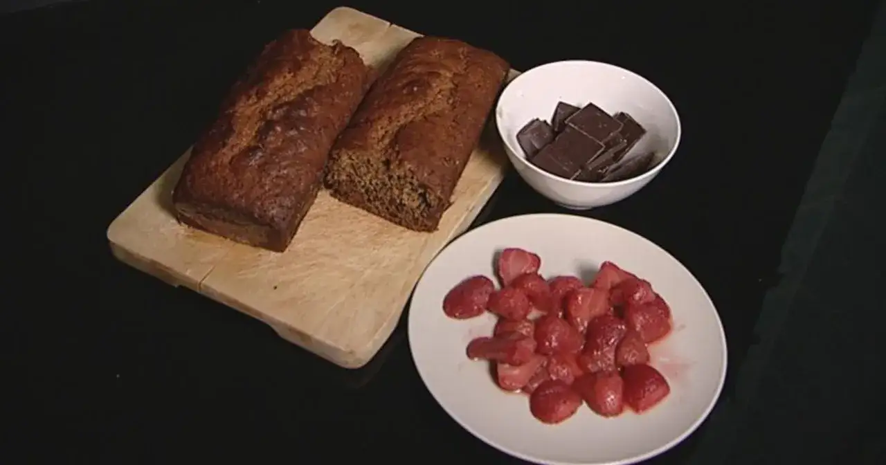 Dwa bochenki chlebka bananowego z bakaliami, obok talerz z truskawkami i miseczka z kawałkami gorzkiej czekolady.