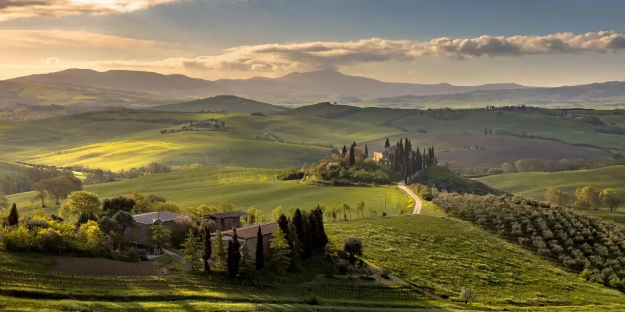 Dolci colline verdi delle Crete Senesi, con un casale e cipressi su una collina, illuminate dalla luce dorata del tramonto.