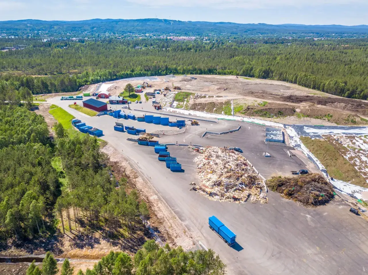 Stor deponi avfall med bl&aring; containrar, en h&ouml;g med tr&auml;avfall och maskiner i arbete.