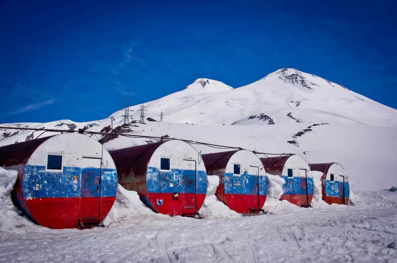 Białe beczki z rosyjską flagą, zasypane śniegiem, pod ośnieżonym szczytem Elbrus.