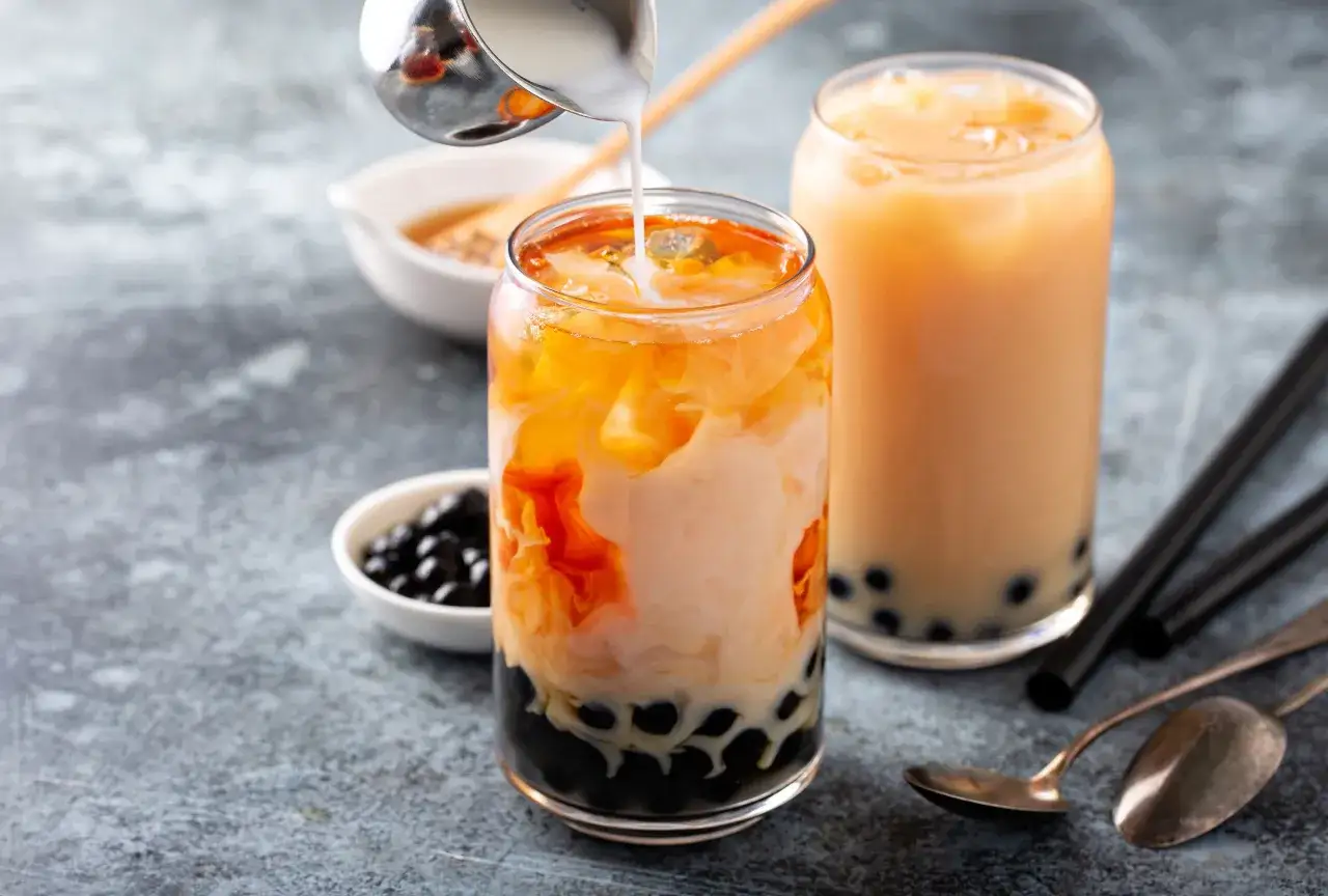 Jak zrobić bubble tea? Mleko wlewane do szklanki z pomarańczowym napojem i czarnymi kuleczkami. Obok drugi nap&oacute;j i akcesoria.