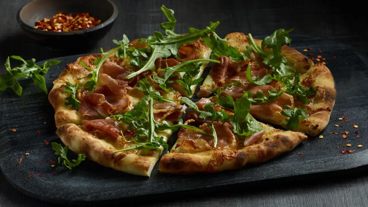 Pyszna pizza prosciutto z rukolą i płatkami chili na ciemnej desce.