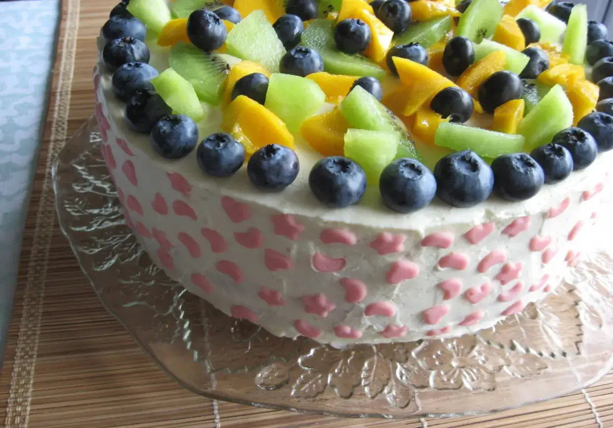Kolorowy tort ozdobiony owocami: borówkami, kiwi i mango. Boki tortu pokryte są białym kremem z lukrem z mascarpone i różowymi ozdobami.