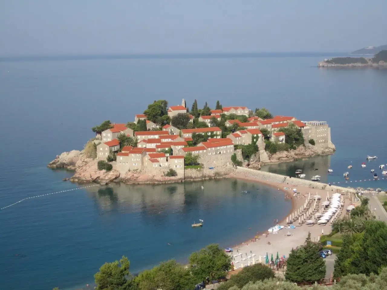 Sveti Stefan, en pittoresk ö med röda tak, ansluten till fastlandet av en smal landremsa. En strand med parasoller och badande syns vid sidan.