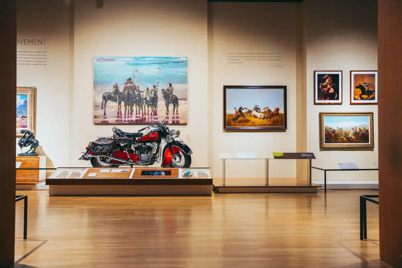 A museum exhibit showcasing western artists, featuring paintings of cowboys and Native Americans, a sculpture of a bucking bronco, and a vintage motorcycle.