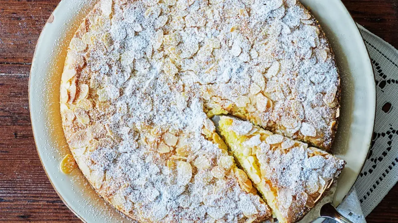 En saftig mandelkaka med citron, toppad med mandelspån och pudersocker. En bit är uppskuren.