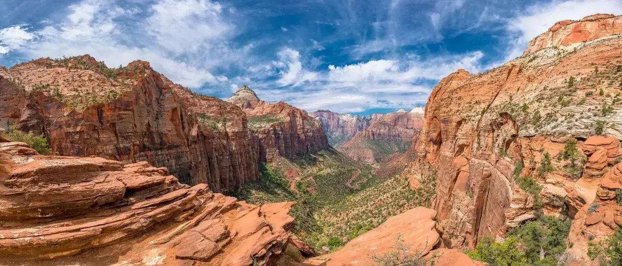 Majest&auml;tische rote Felsformationen im Zion Nationalpark unter blauem Himmel mit wei&szlig;en Wolken. Ein tiefer Canyon mit gr&uuml;ner Vegetation.