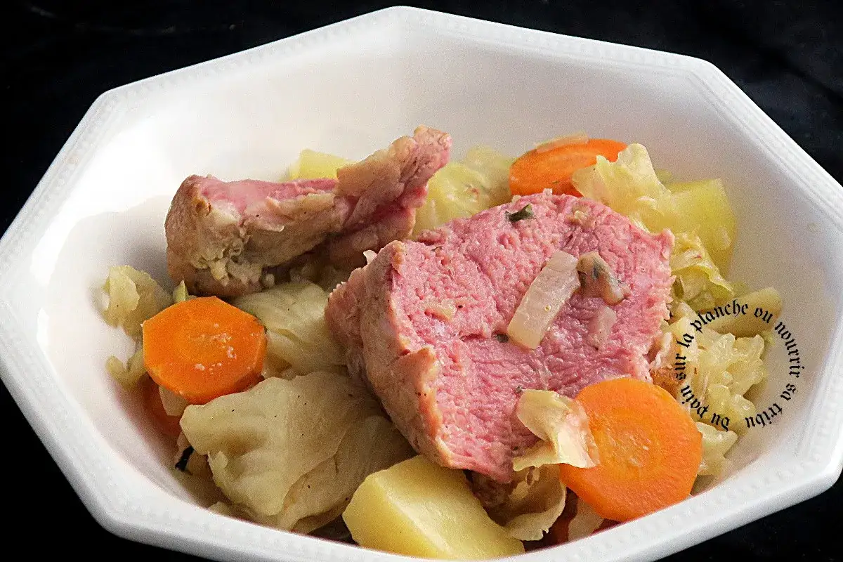 Une g&eacute;n&eacute;reuse pot&eacute;e au chou, avec du porc rose, des carottes et des pommes de terre, servie dans un plat blanc.