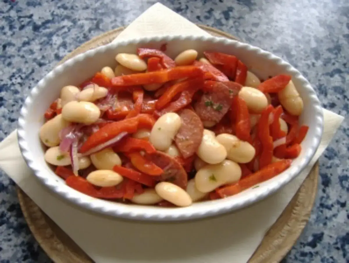 Ein herzhafter spanischer Bohnensalat mit weißen Bohnen, roten Paprikastreifen und Wurstscheiben in einer weißen Schüssel.